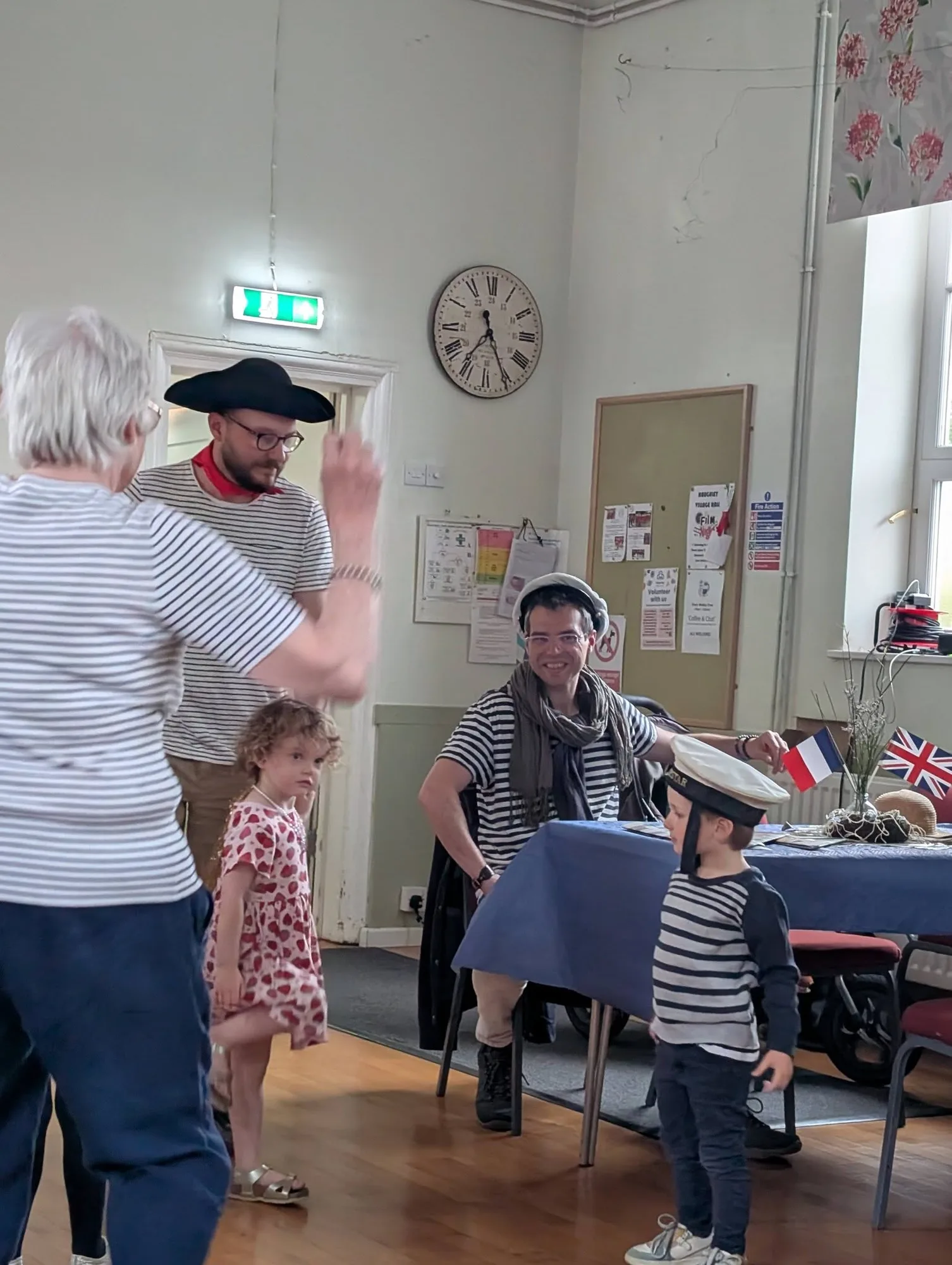 Two adults in striped Breton tops and berets engage with two young children during a French-themed HAWTA community event, with a French flag and Union Jack visible on a table in the background.